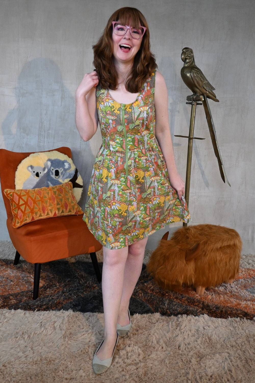 Small brown haired model in glasses and scoop neck sleeveless olive orange and green mushroom and flower print dress