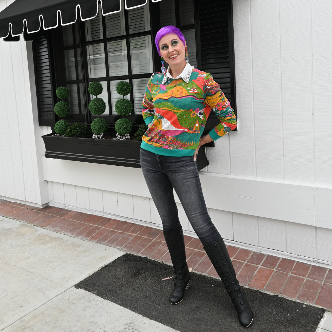 Full body view of purple-haired model wearing landscape print sweatshirt and standing in front of a window 