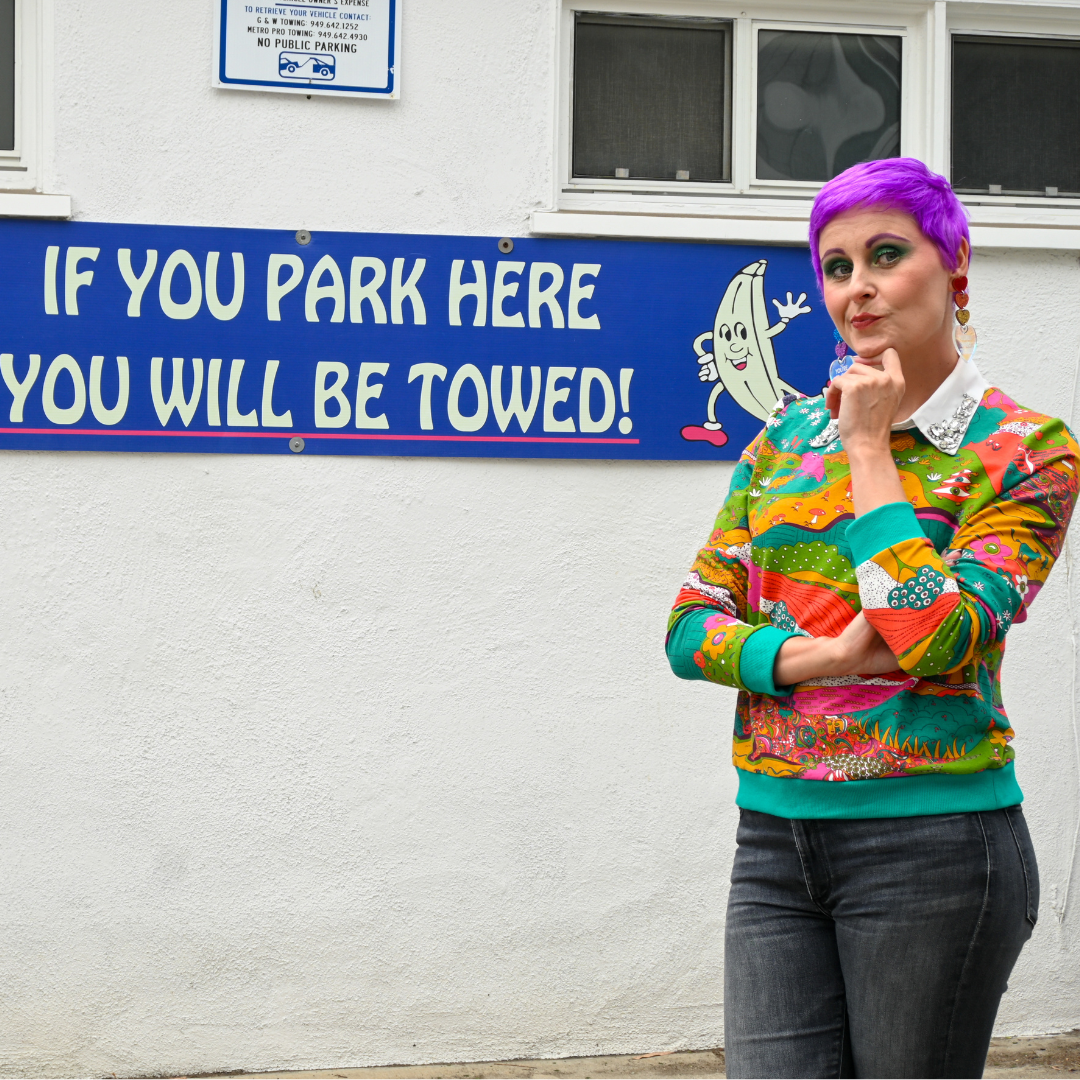 Purple-haired model in bright, multi-colored sweatshirt and grey jeans