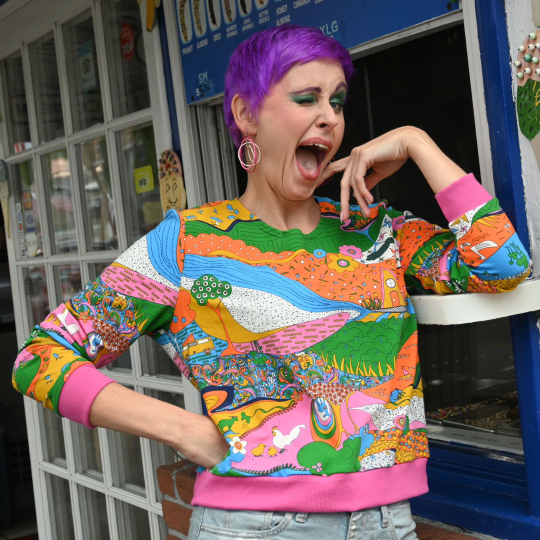 Purple-haired model in rainbow printed sweatshirt and jeans, winking