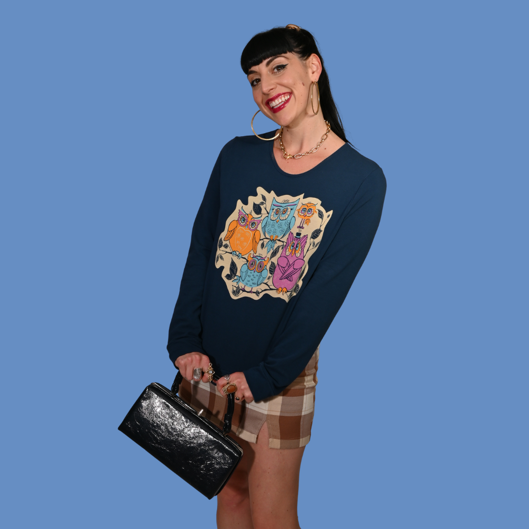 Woman wearing a navy blue sweatshirt with a colorful owl design, holding a black clutch against a blue background