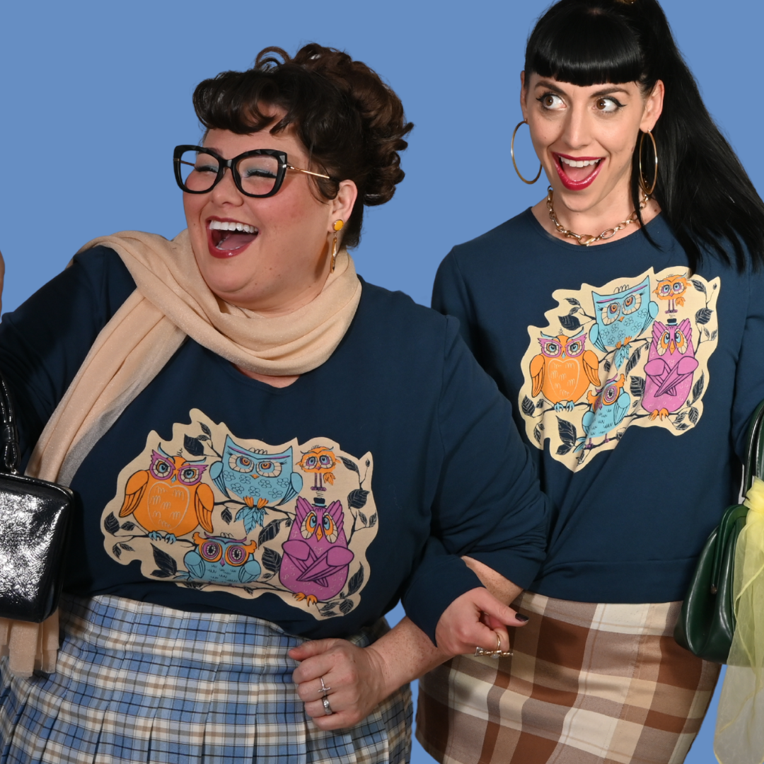 Two women wearing navy blue sweatshirts with colorful owl designs against a blue background.