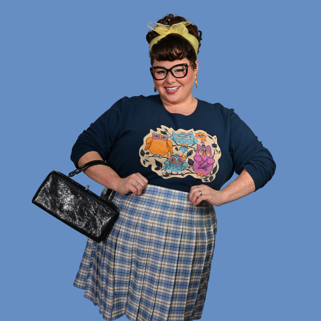 Person wearing a navy sweatshirt with a colorful graphic design, plaid skirt, and holding a black handbag against a blue background.