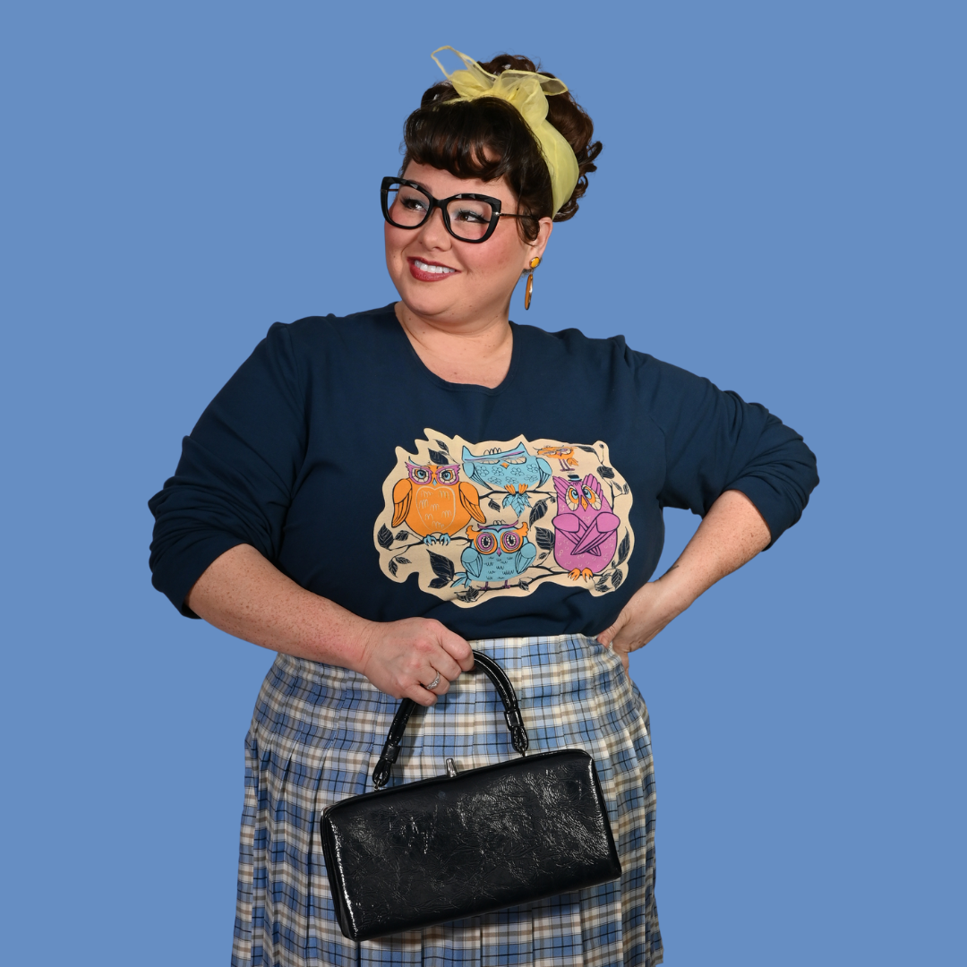 Pretty woman wearing a navy sweatshirt with cartoon owls and a plaid skirt, holding a black handbag against a blue background.