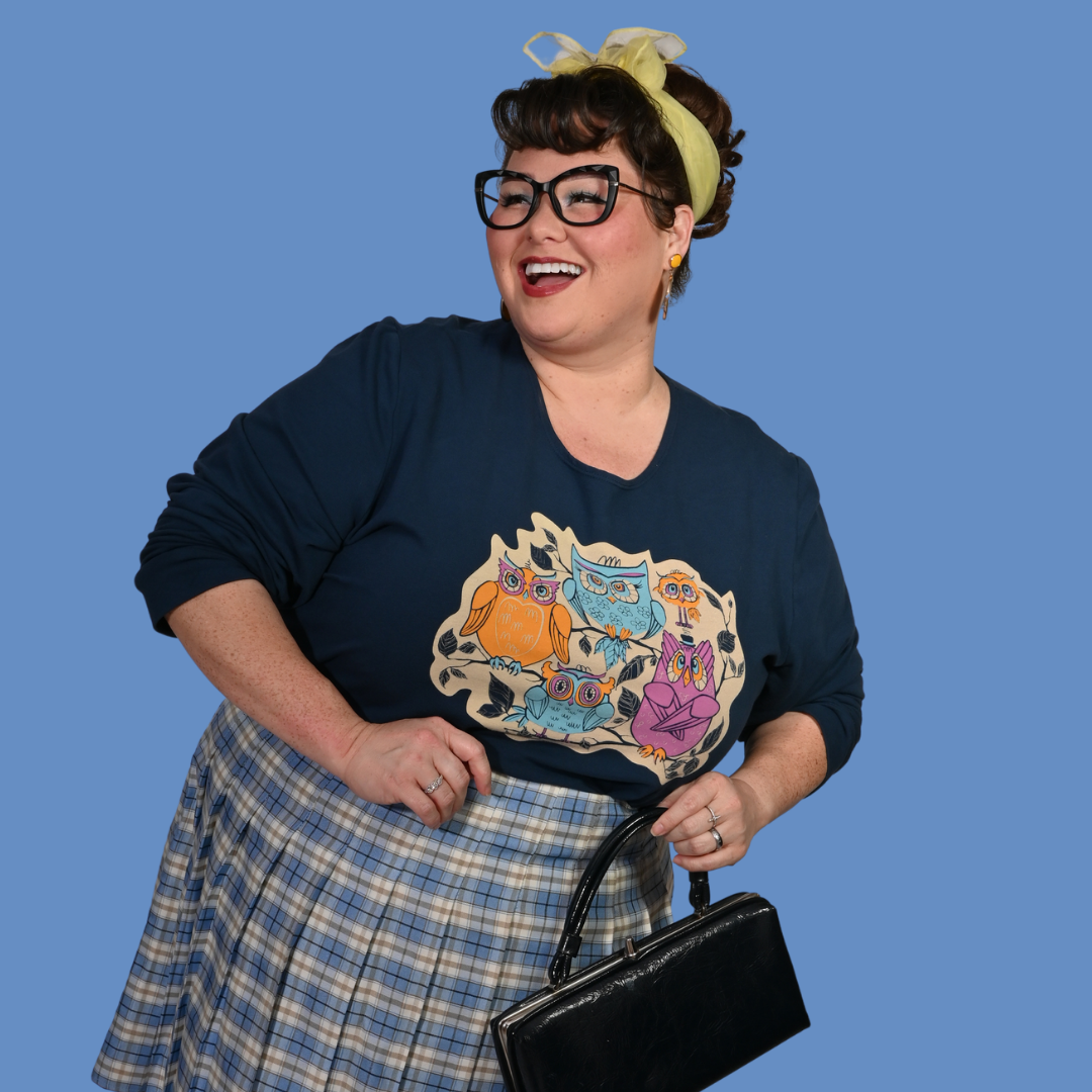 Dark-haired woman wearing a navy blue shirt with a graphic design, plaid skirt, and holding a black handbag against a blue background.