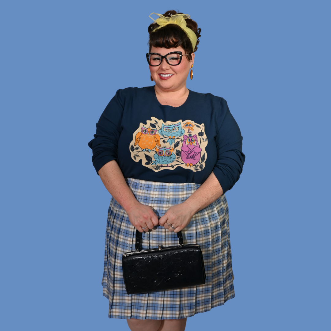 Person wearing a navy sweatshirt with colorful owls, plaid skirt, and holding a black handbag against a blue background.