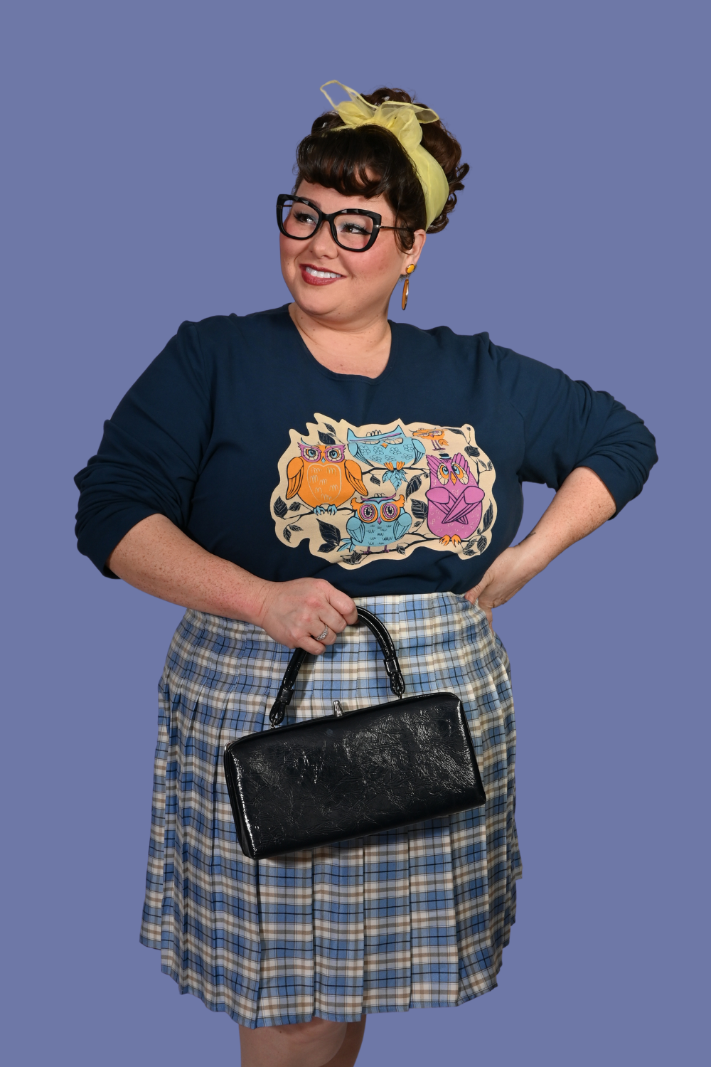 Person wearing a navy sweatshirt with a colorful graphic design of owls and a plaid skirt, holding a black handbag on a blue background.