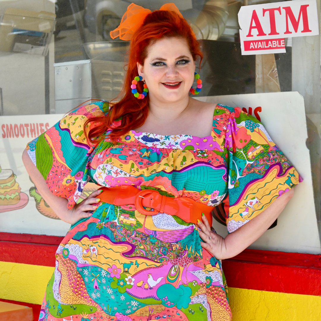 Closeup of red-haired model in brightly colored printed dress and fun accessories, in front of donut shop