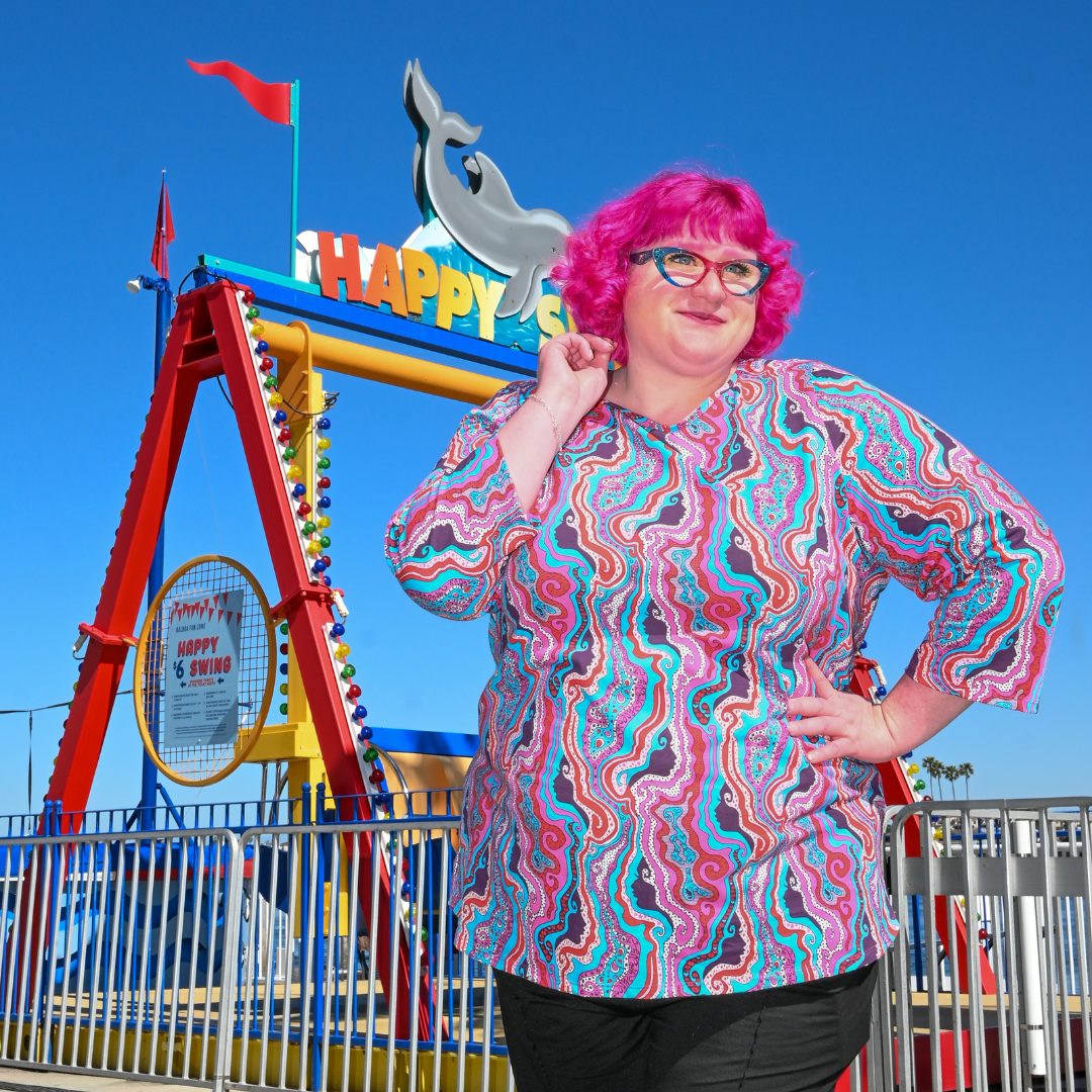 Pink-haired model in swirling psychedelic print tee in front of kids' ride