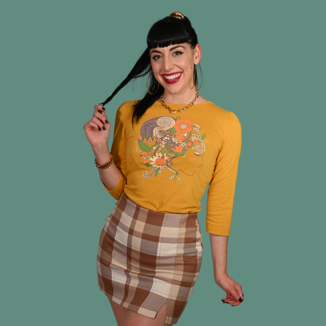 Woman wearing a mustard yellow graphic tee with a colorful design and a plaid skirt, posing against a green background.