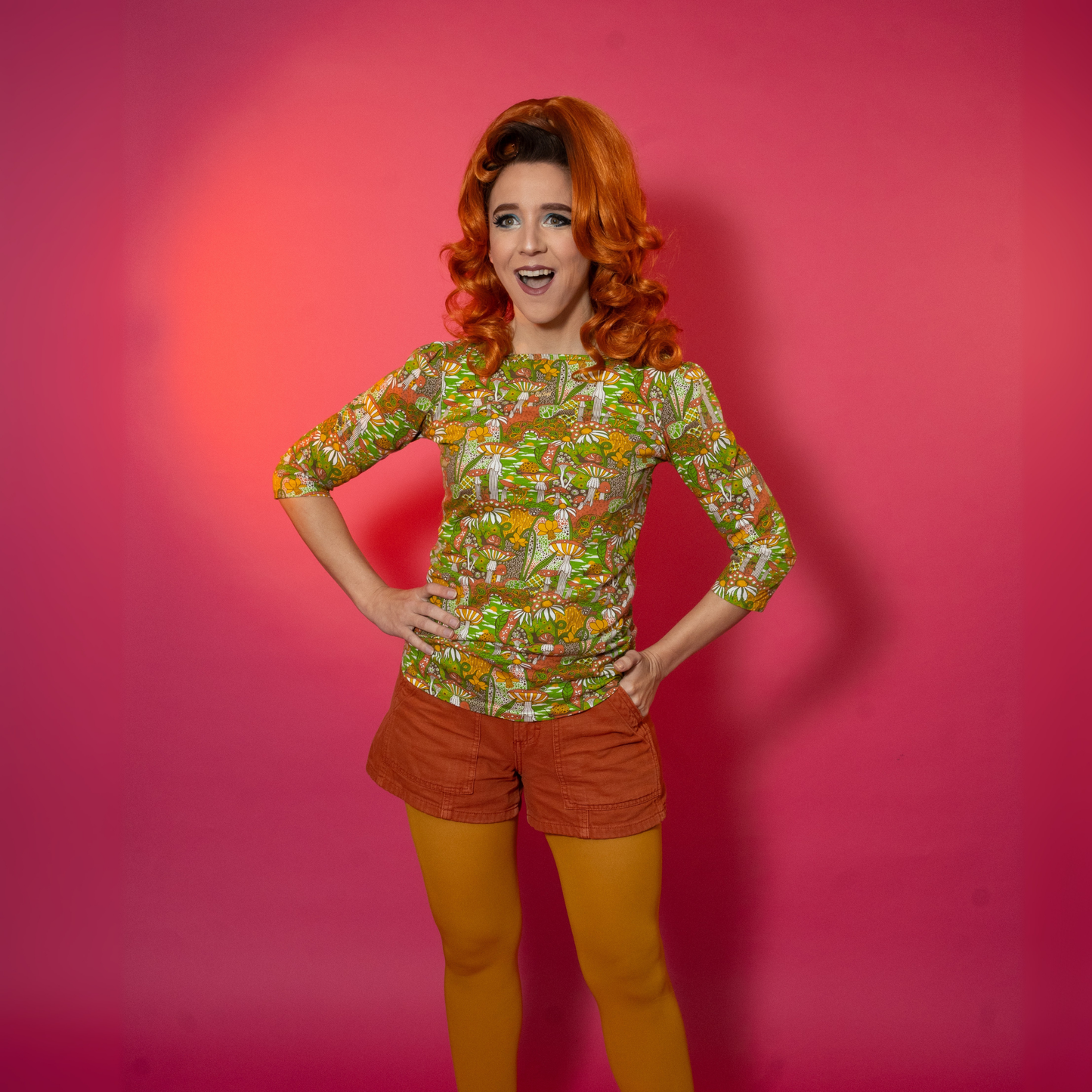 Person wearing a patterned shirt and orange shorts and yellow tights against a pink background