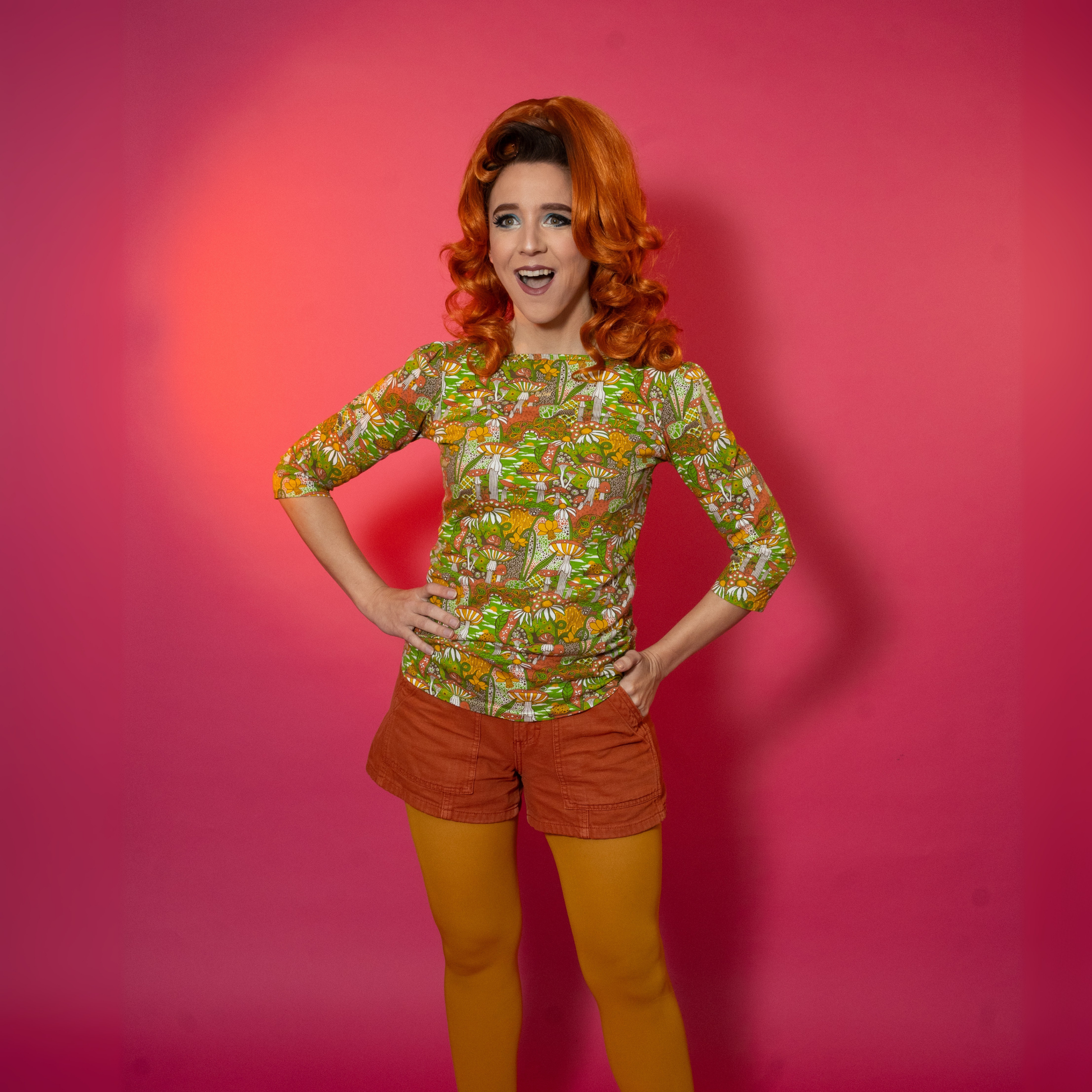 Person wearing a patterned shirt and orange shorts and yellow tights against a pink background