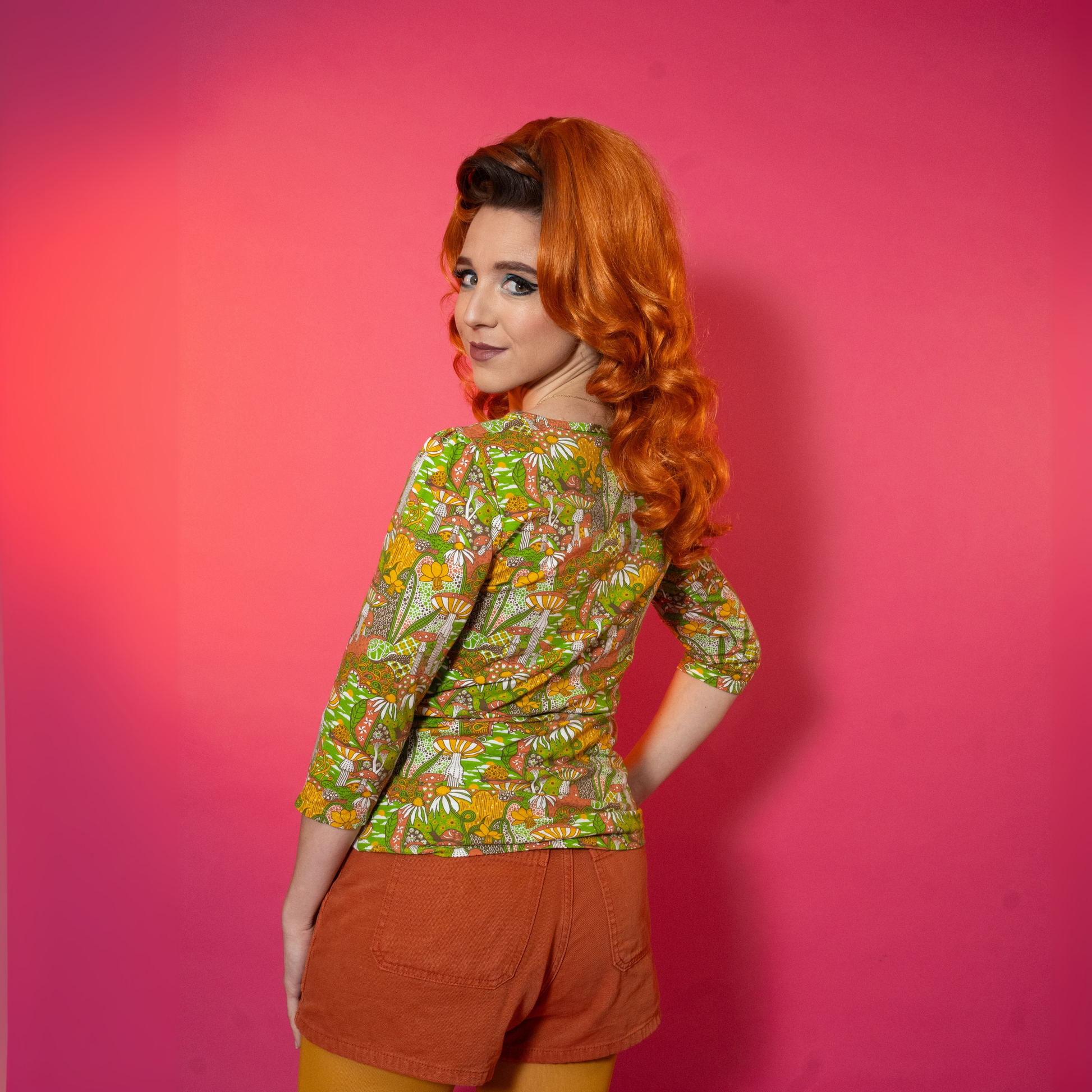 Back view of woman wearing a floral top and orange shorts against a pink background