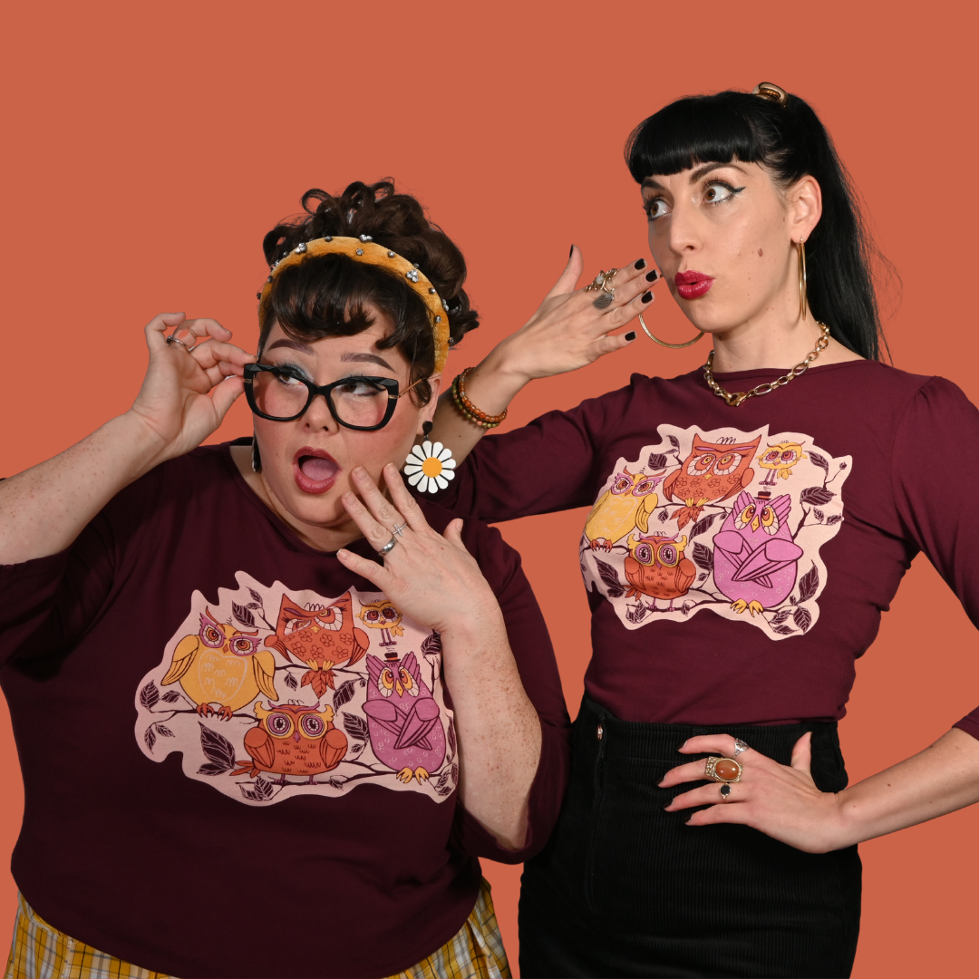 Closeup of two women wearing reddish purple plum t-shirts with owl designs on an orange background