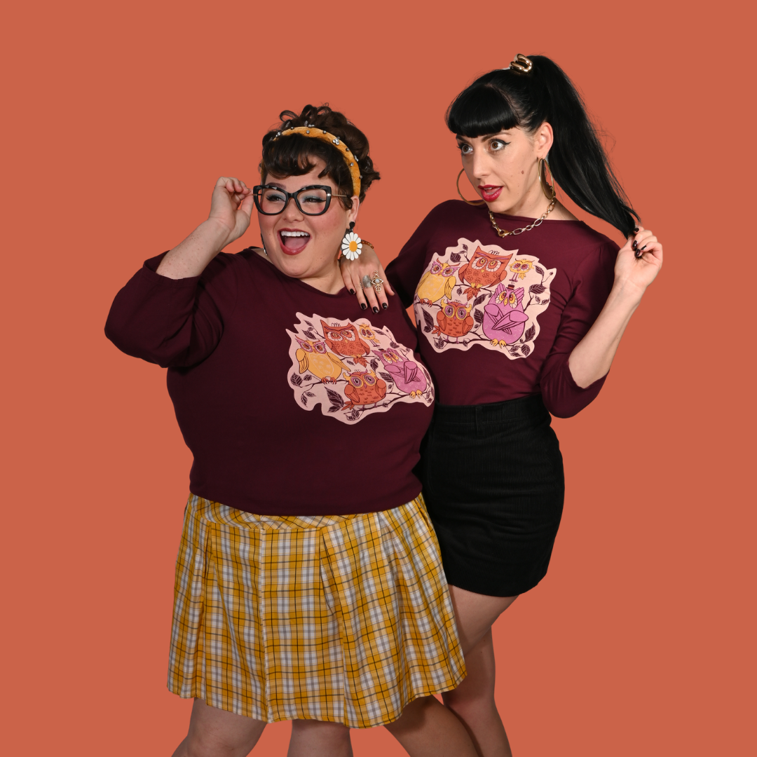 Two women wearing plum purple graphic tees with colorful vintage owl designs against an orange background