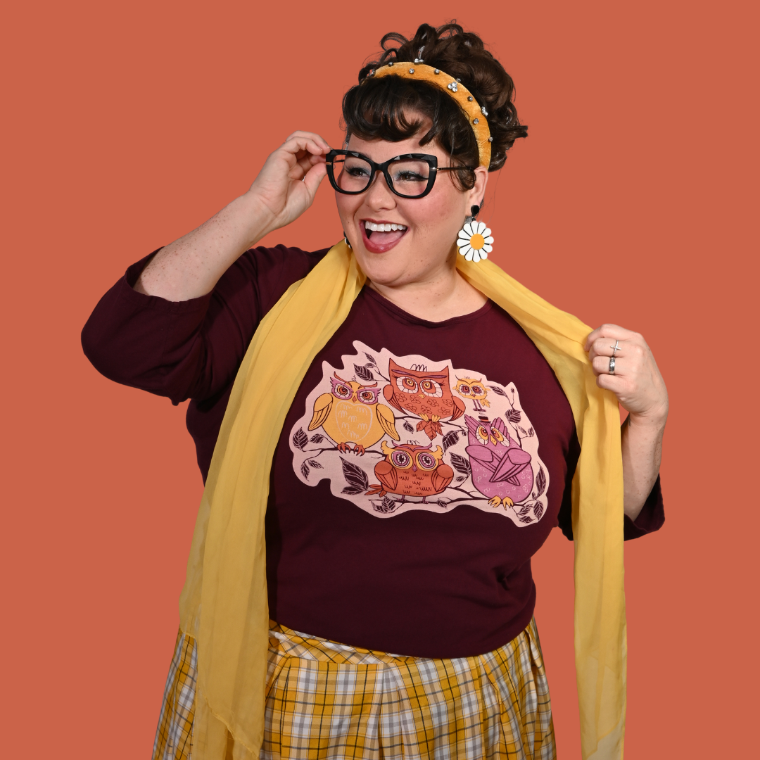 Person wearing a dark purple shirt with owl design, yellow scarf, and plaid skirt on an orange background