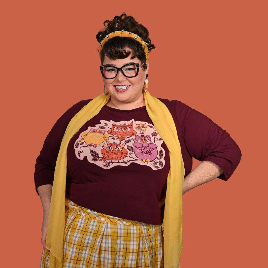 Person wearing a red purple graphic tee with owl design, yellow scarf, and plaid skirt on an orange background