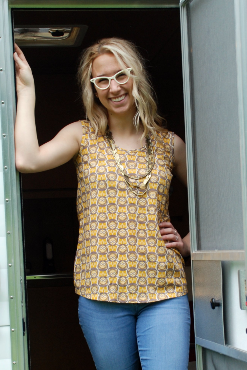 Cute girl wearing glasses and sleeveless yellow and grey girl print top, standing in the door of a camper