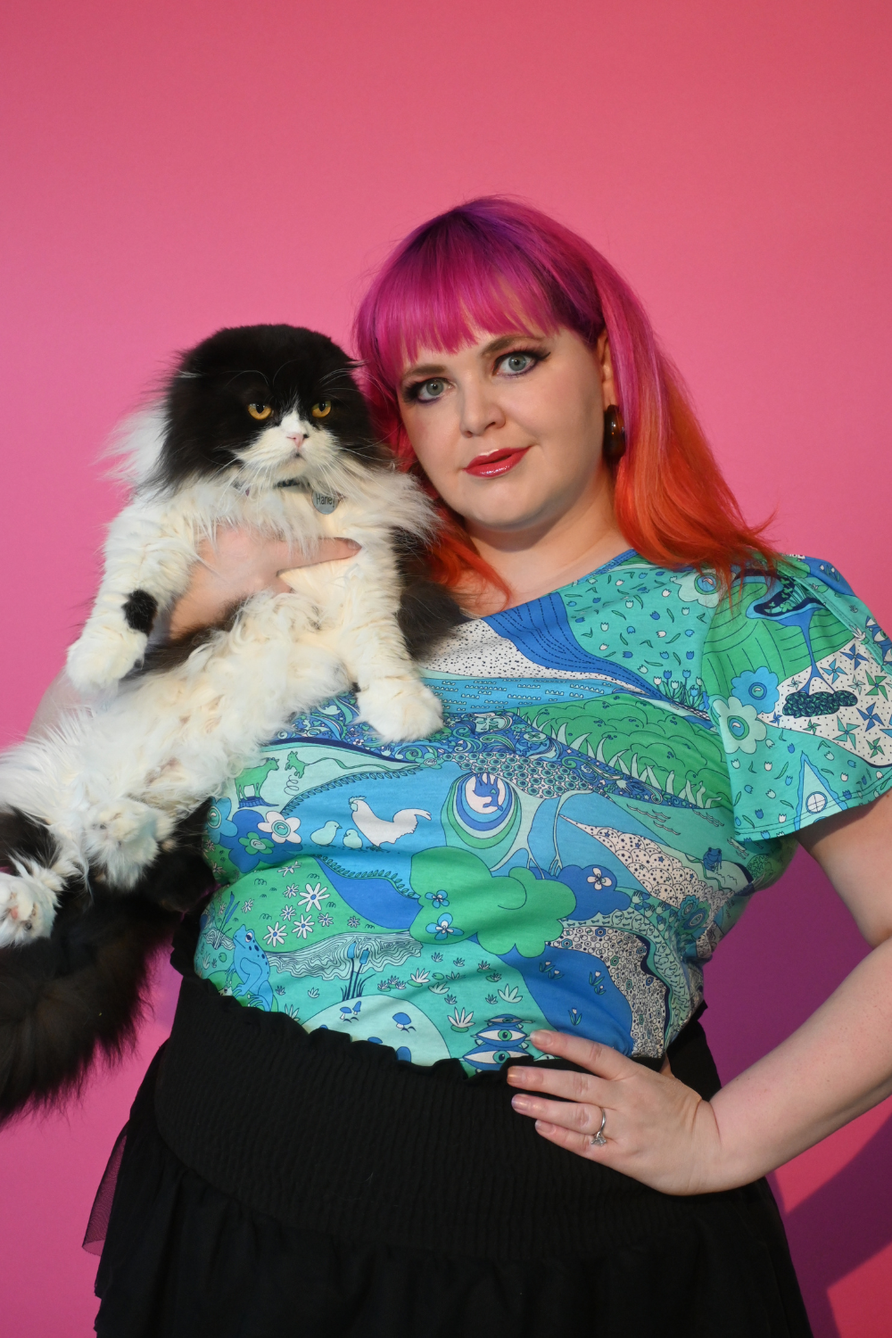 Pink haired model holding cat wearing shirt with landscape graphic in green and blue
