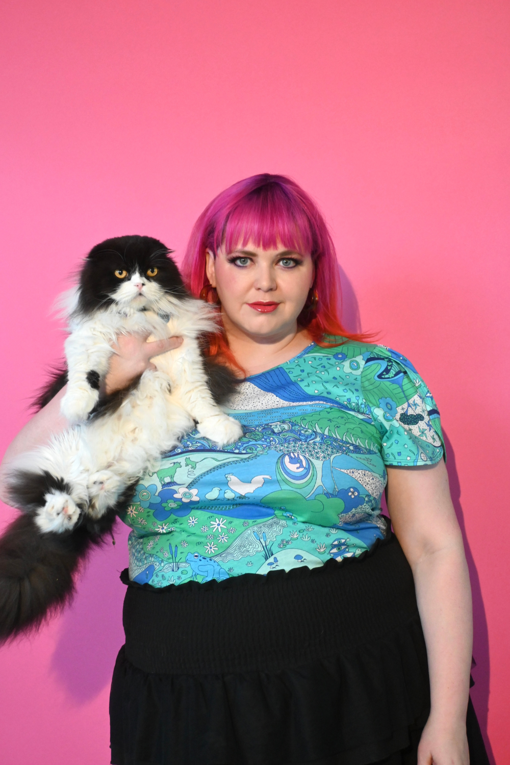 Pink haired model holding cat wearing shirt with landscape graphic in green and blue