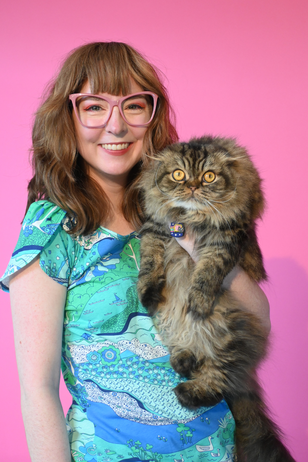 Model wearing glasses and holding cat wearing shirt with landscape graphic in green and blue