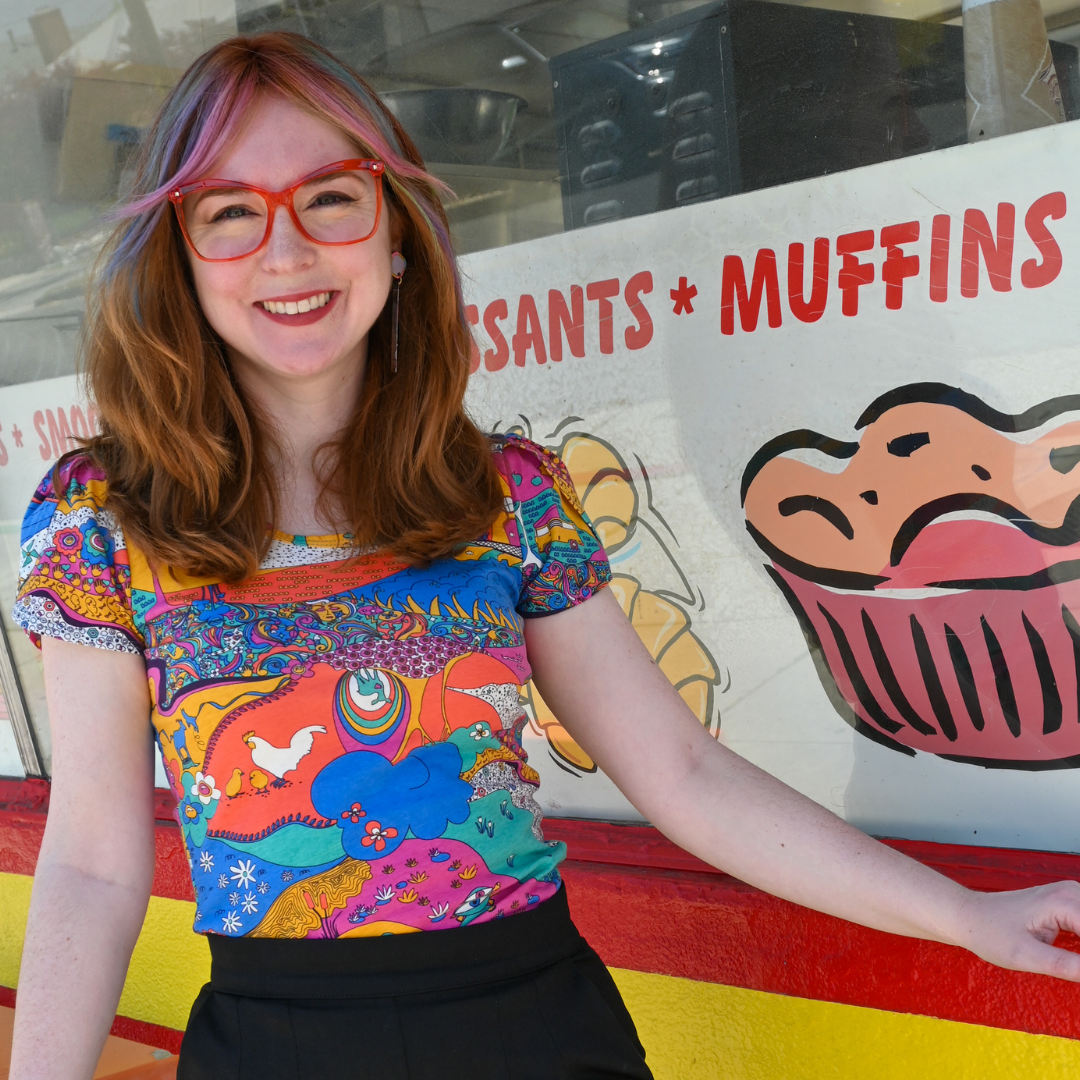 Smiling model in square glasses and a brightly colored tulip sleeved tee and shorts in front of a bakery