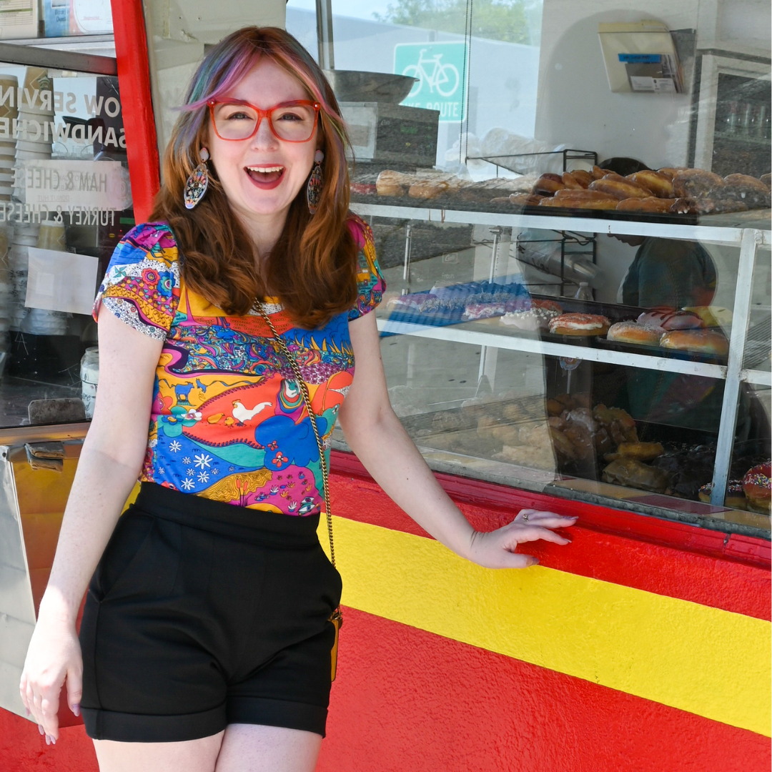 A cute model in glasses, a brightly colored printed tee, and black shorts in front of a donut counter