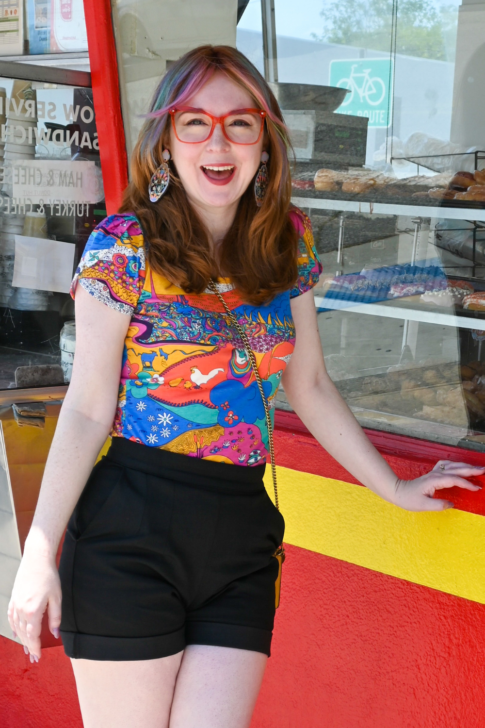 Smiling model wearing brightly colored printed tee, shorts, and red glasses