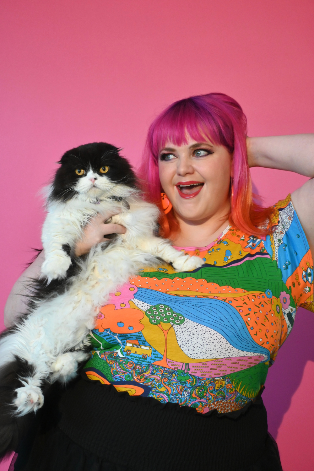 Pink haired model holding cat wearing shirt with landscape graphic in multicolor