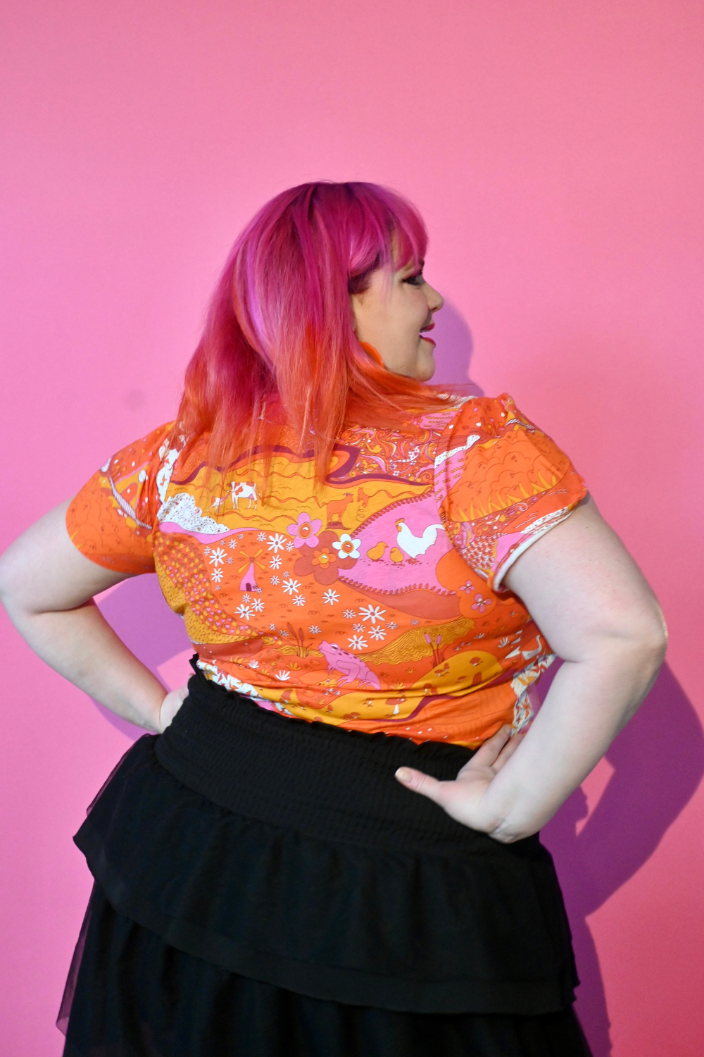 Back view of pink haired model wearing shirt with graphic of a landscape in orange and pink