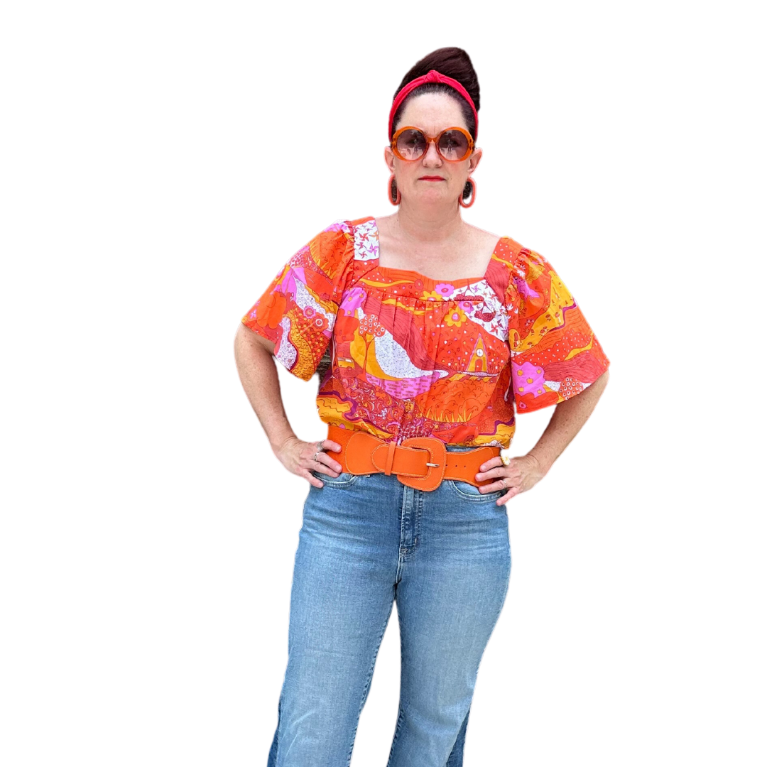 Model in brightly colored woven square-neck top with landscape print in pink, orange and yellow, tucked into jeans