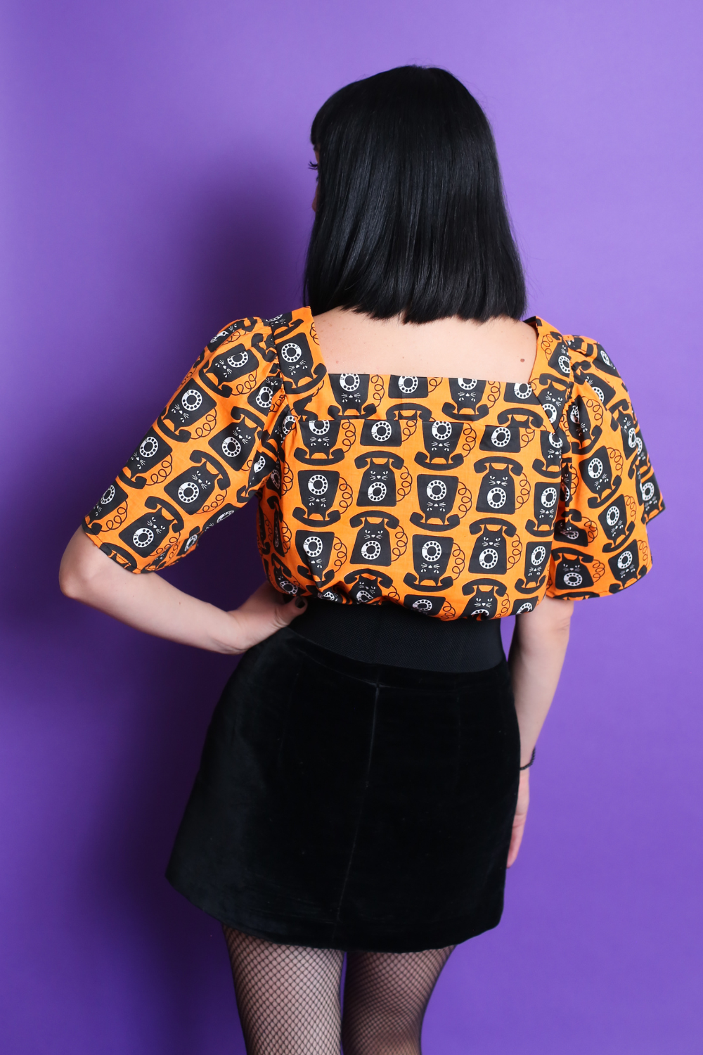 Black haired model in orange and black cat phone print top & mini skirt, back view