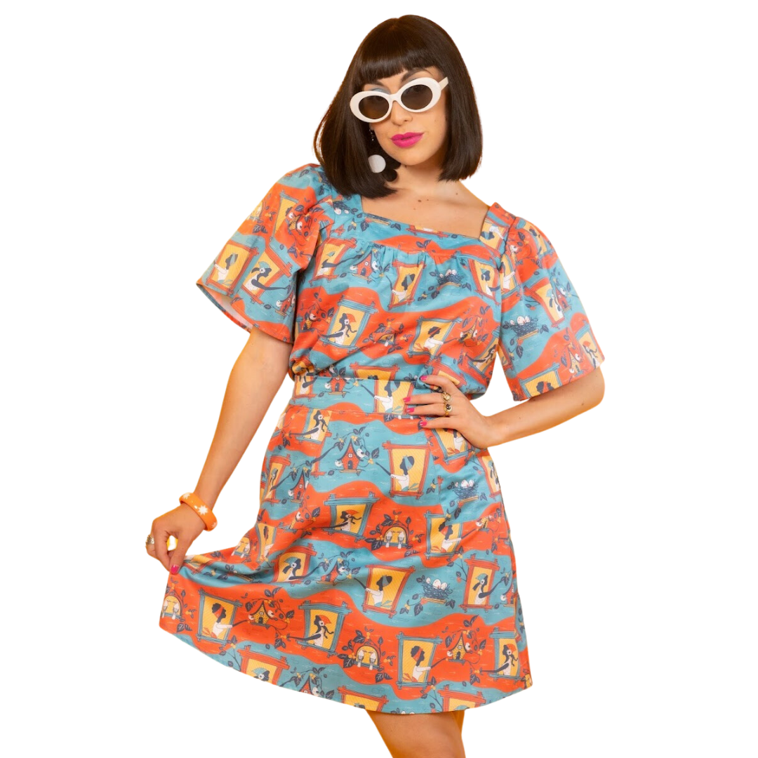 Dark-haired model in matching skirt and shirt with red, yellow and blue graphic print of girls and birdhouses