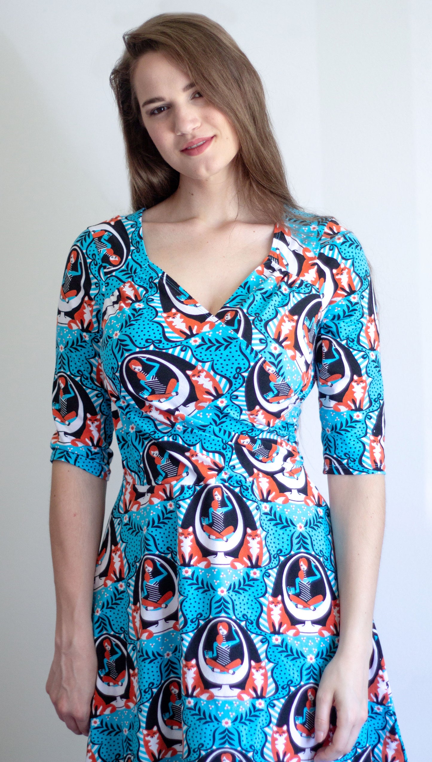 Model in blue sleeved wrap dress featuring print of foxes and girls reading books sitting in an egg chair