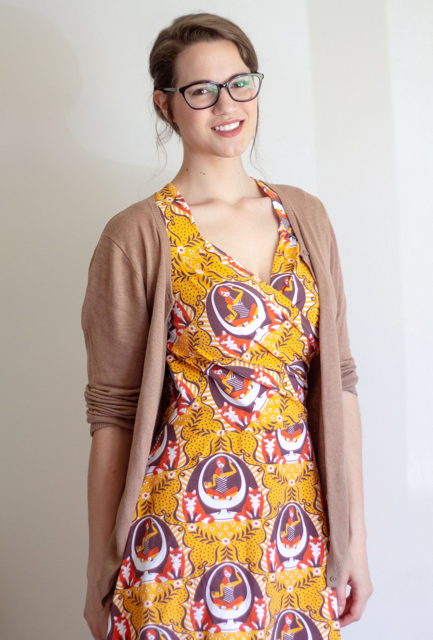 Closeup of model in yellow and orange sleeved wrap dress featuring print of foxes and girls reading books in an egg chair, with sweater