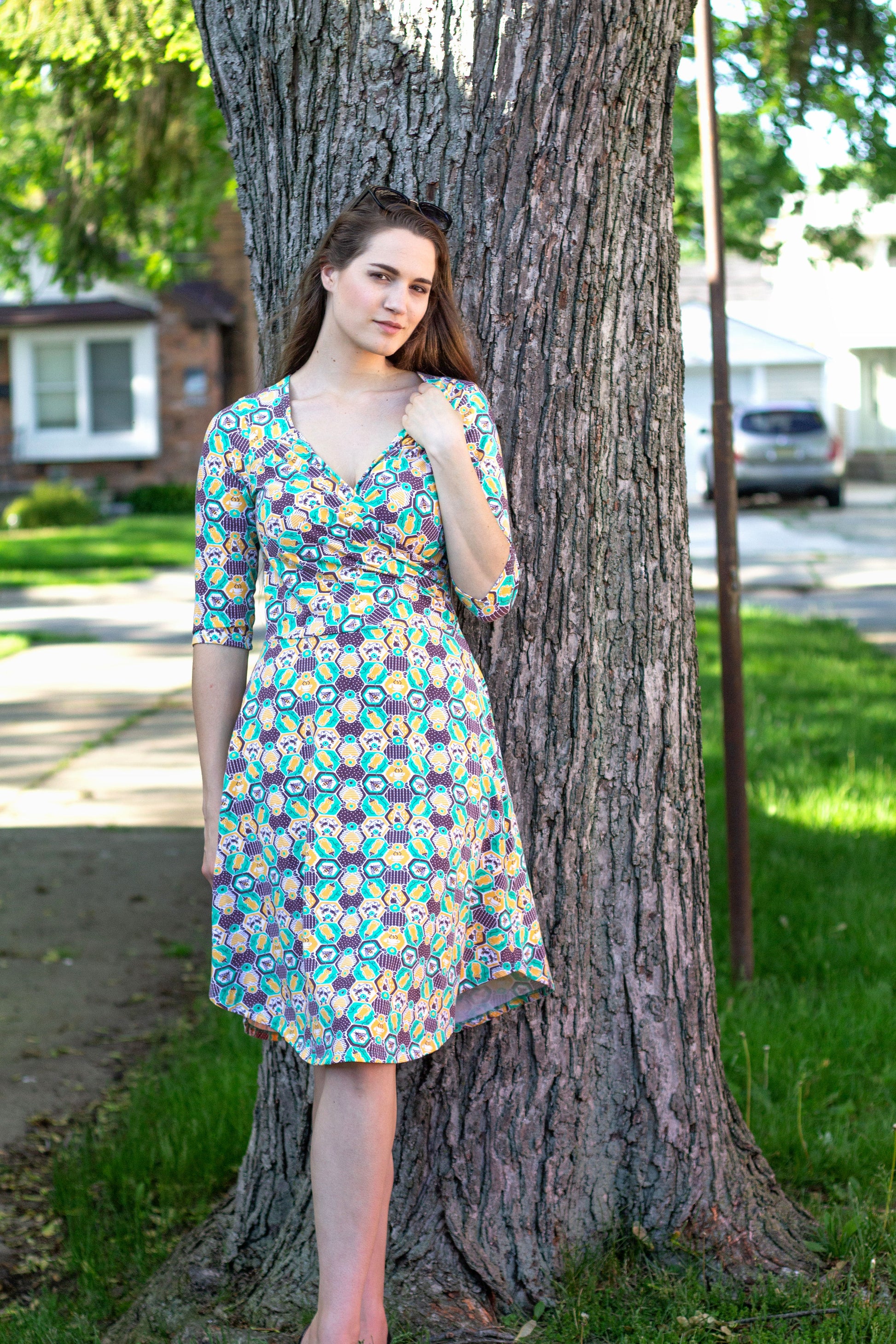 Brown-haired model wearing yellow, green and brown honeybee and hexagon print wrap dress and standing in front of a tree 