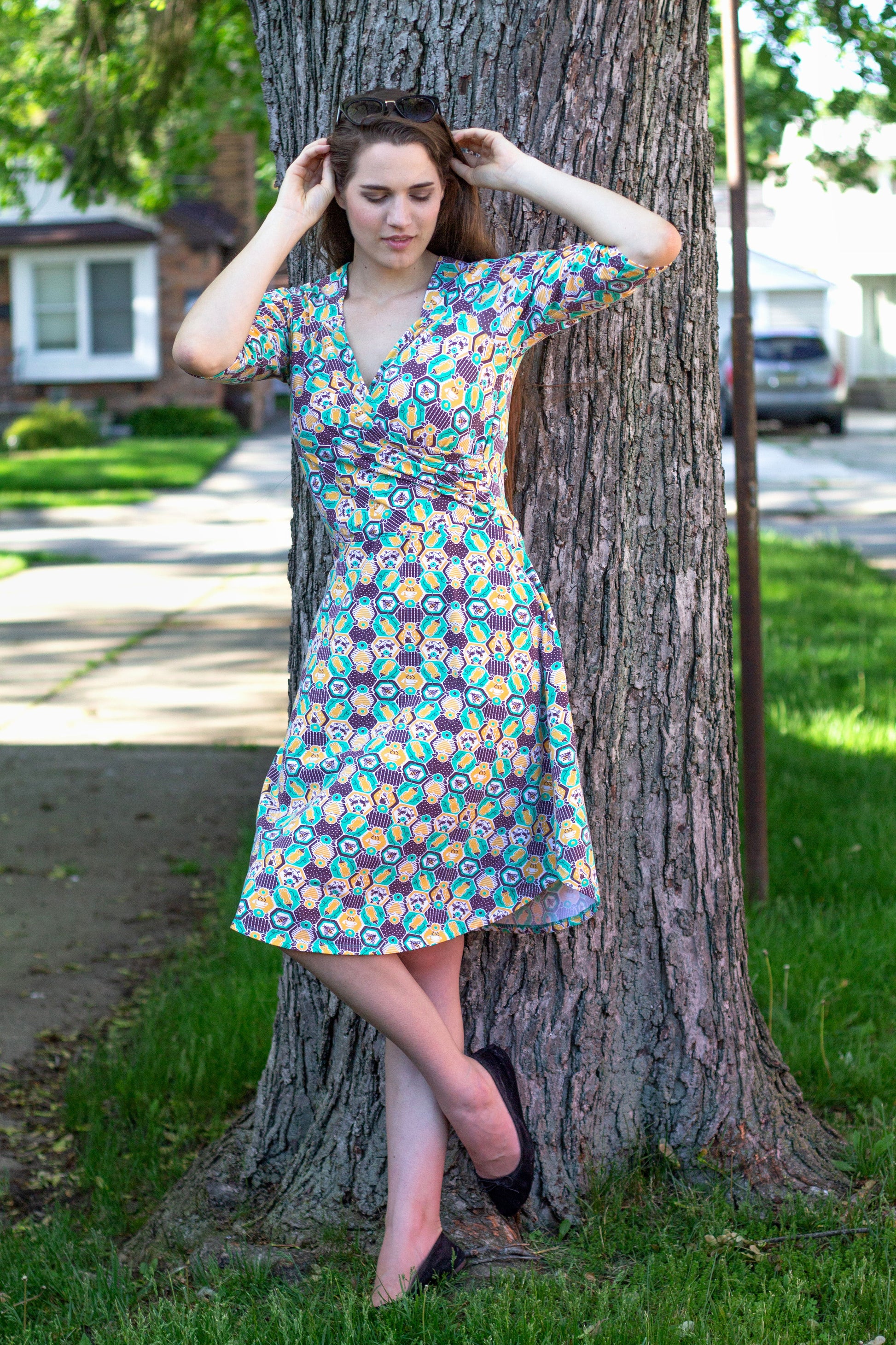 Brown-haired model wearing yellow, green and brown honeybee and hexagon print wrap dress in front of a tree