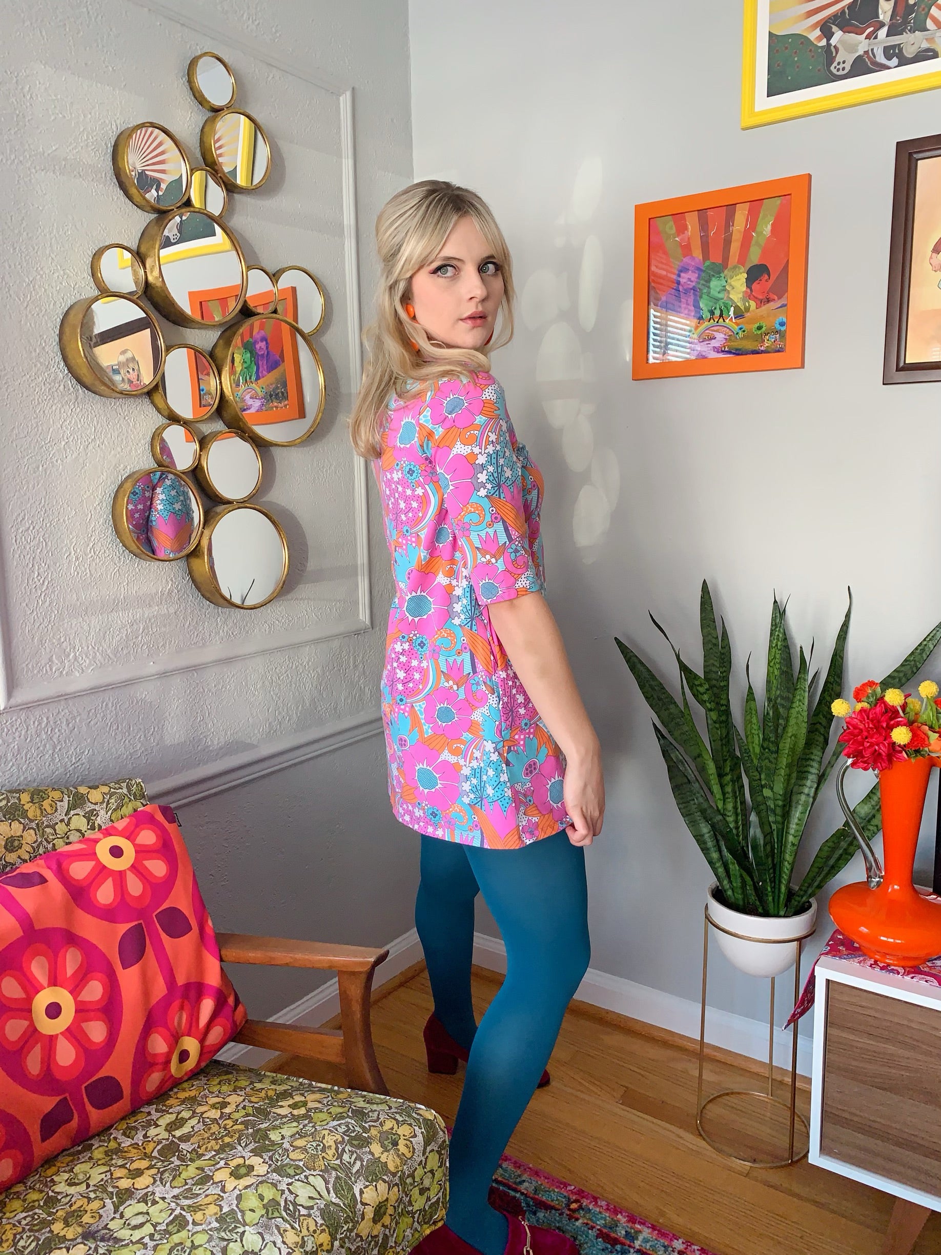 Side view of bright pink, orange and aqua retro floral print a-line pocket tunic on blonde model in cute house