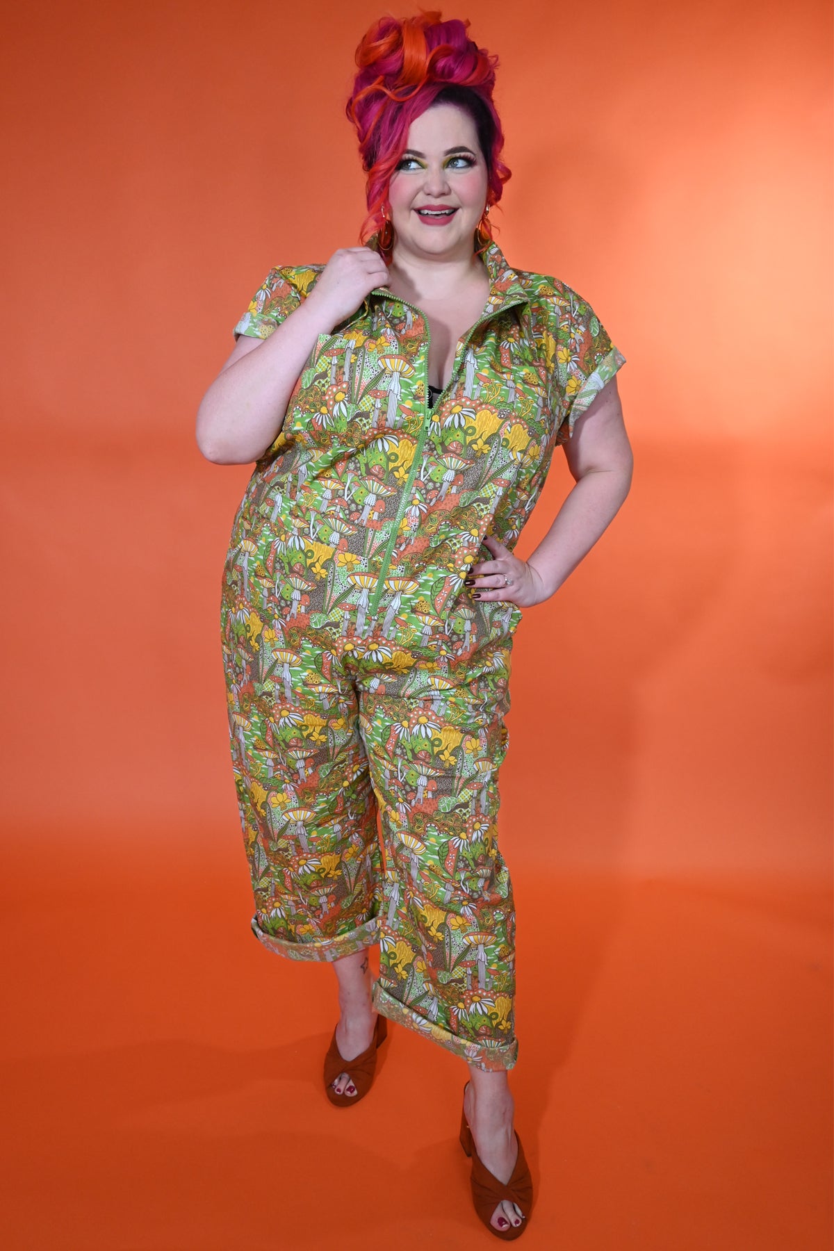 Happy pink-haired model in olive green, orange and yellow mushroom, snail and flower print jumpsuit with big zipper and pockets 