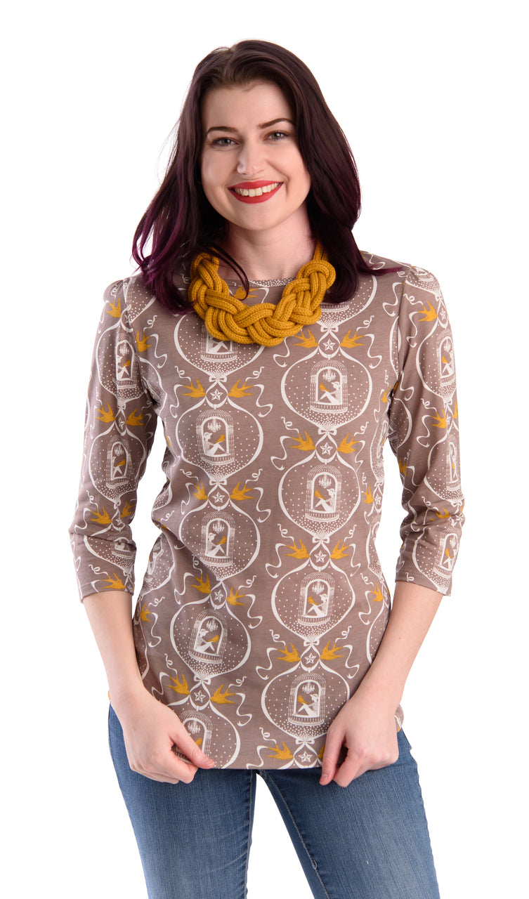 Dark haired model wearing 3/4 sleeve tee in beige with graphic of girl sitting with bird in hand