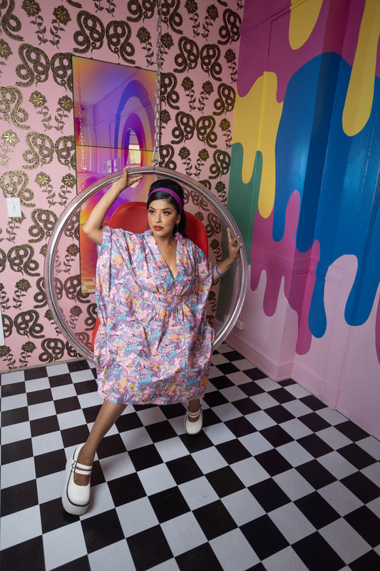 Dark-haired model wearing pink aqua and yellow mushroom tunic and sitting in bubble chair in colorful room of patterns