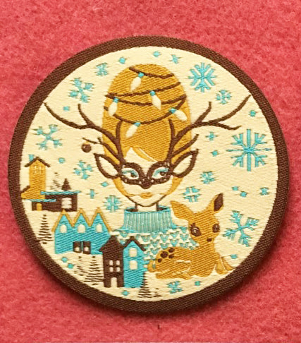 Round iron on patch with brown, gold and aqua print of Christmas girl with beehive hair, deer and putz houses