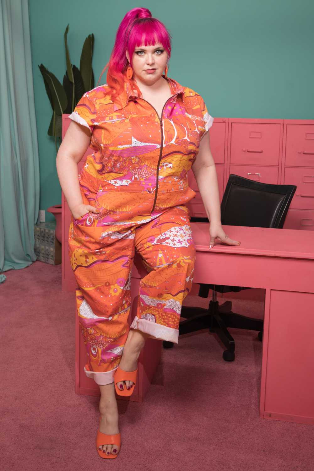 Pink-haired model leaning on office desk while wearing psychedelic jumpsuit in reds, oranges, and pink