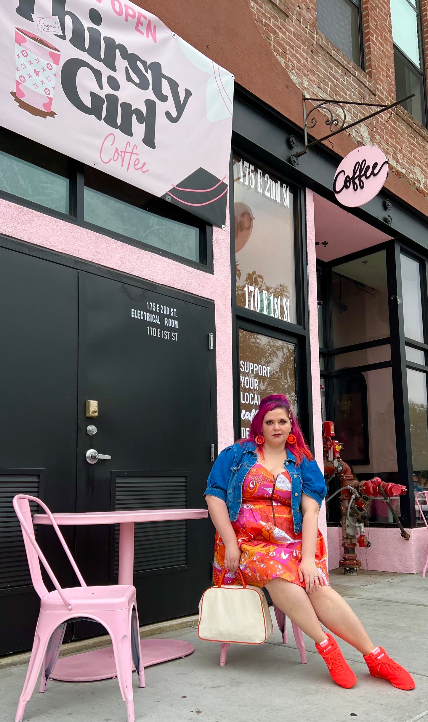 Pretty pink-haired model in bright red orange and pink dress with front zip, jean jacket and red shoes sitting outside a coffee shop