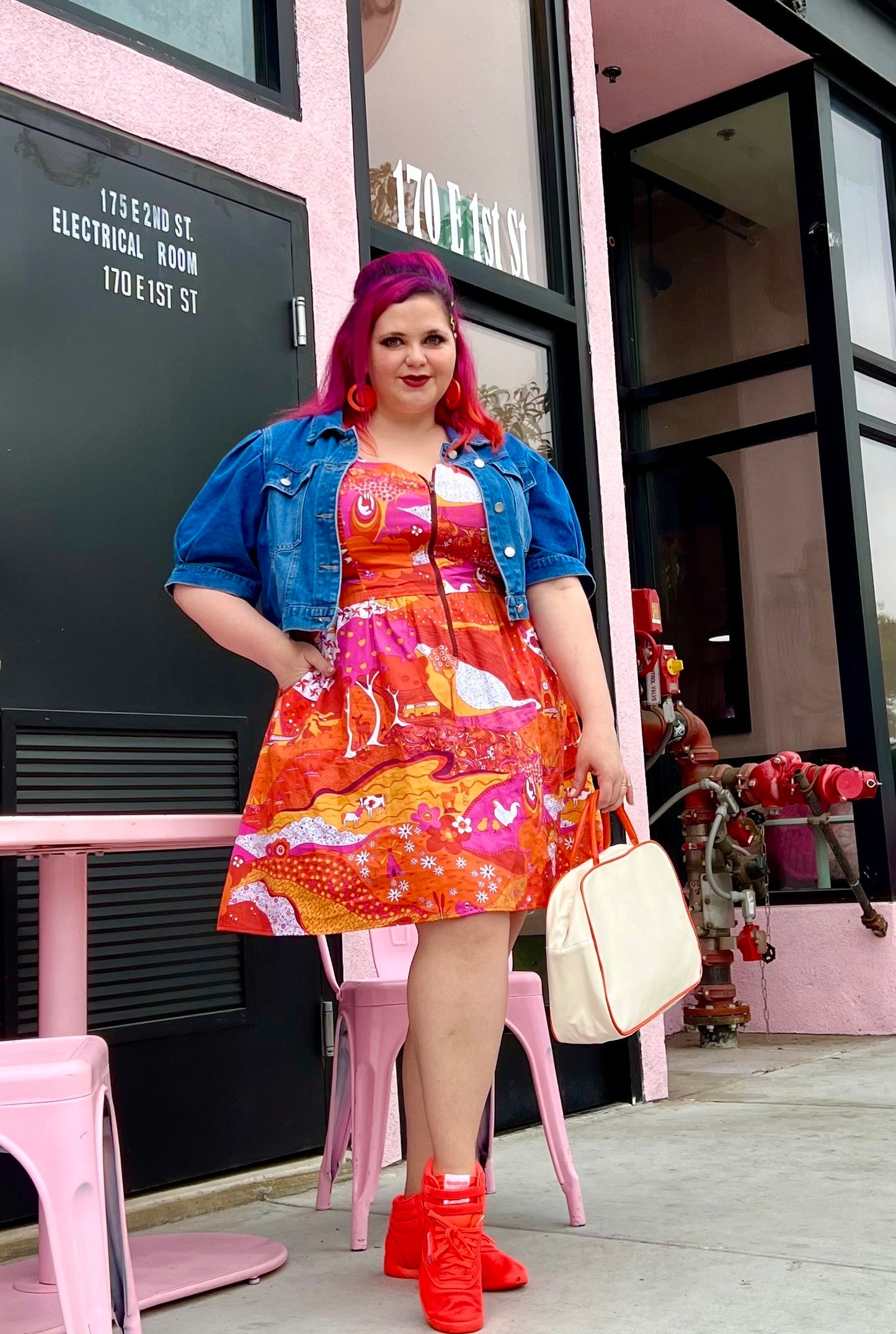 Pretty pink-haired model in bright red orange and pink dress with front zip, jean jacket and red shoes outside a coffee shop