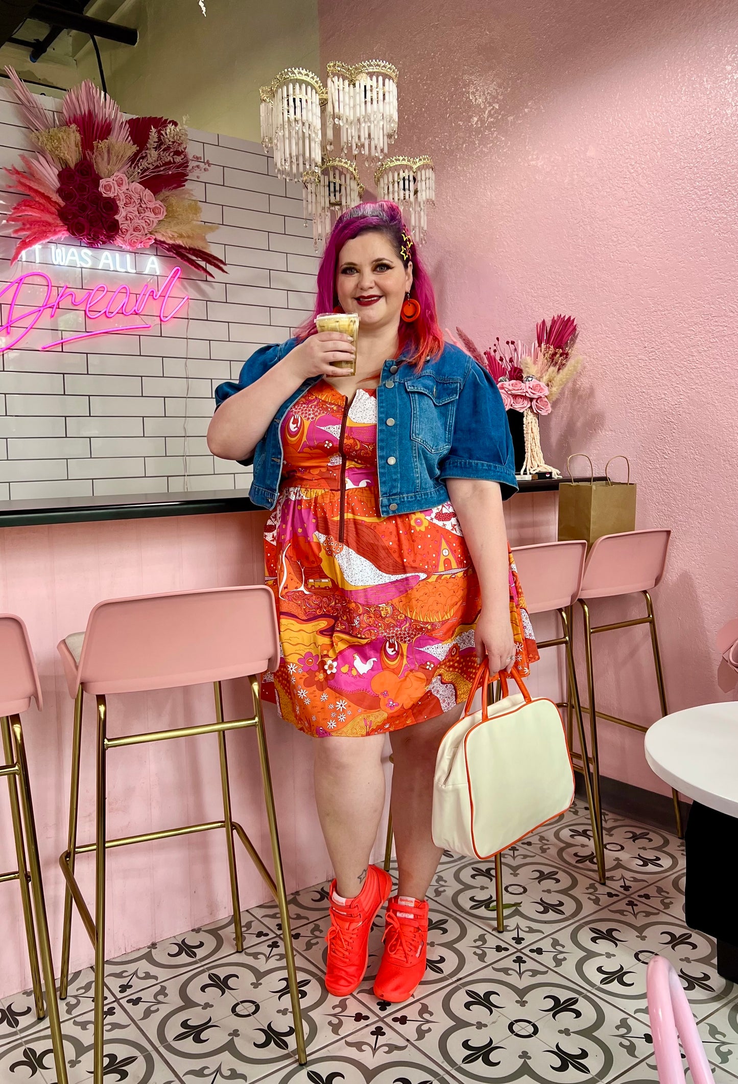 Pretty pink-haired model in bright red orange and pink dress with front zip, jean jacket and red shoes in a coffee shop