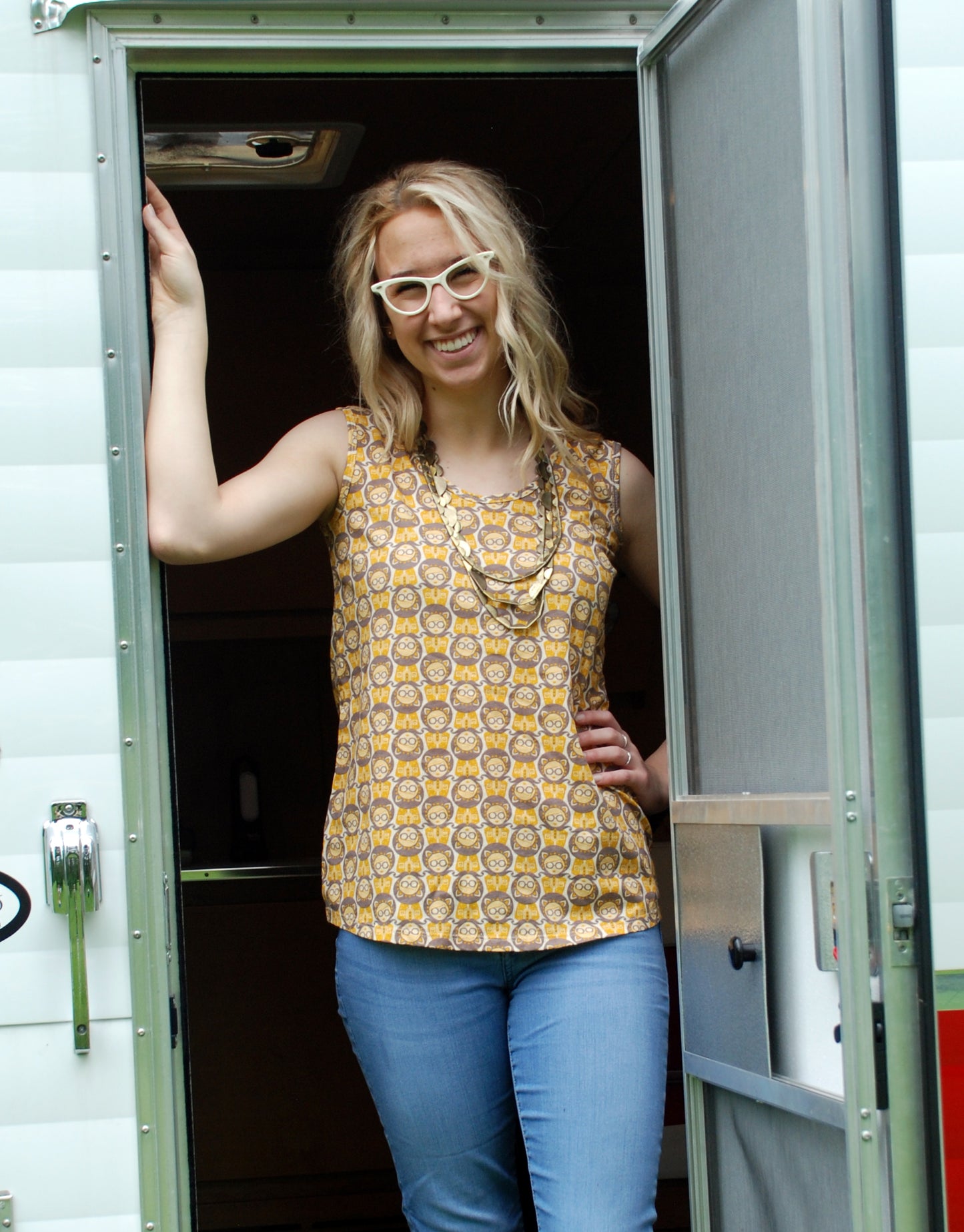 Cute girl wearing glasses and sleeveless yellow and grey girl print top, standing in the door of a camper