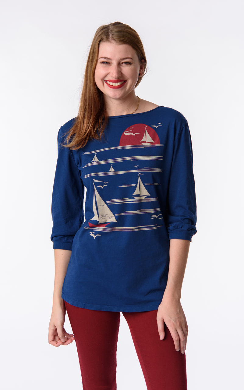 Blonde model wearing long sleeve blue shirt with graphic of boats on the water at sunset