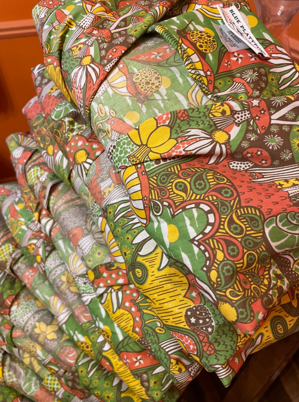 A stack of v-neck olive green, yellow, and orange mushroom, floral and snail print midi caftans