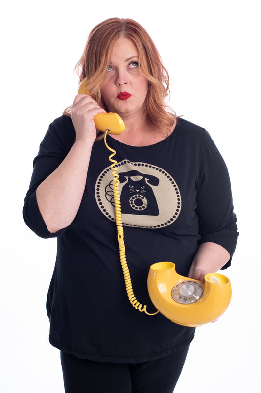 Red haired plus size model talking on donut phone while wearing black cat phone graphic 3/4 sleeve tee and with polka dot background